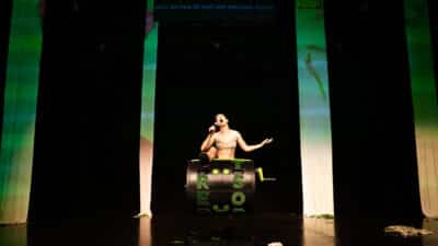 A person in cream-coloured underwear and an open green button up shirt, with gold gloves, arms in the air, standing on a black stage