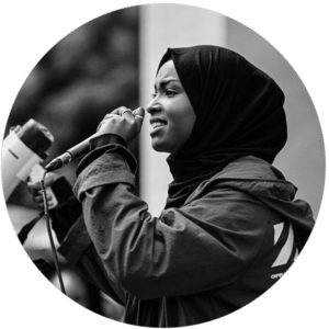 A young woman wearing a hijab and coat is holding a loudspeaker, speaking into a microphone