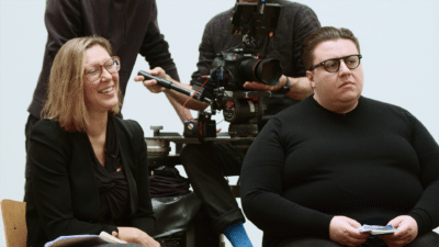 Two people sit with a camera crew behind them